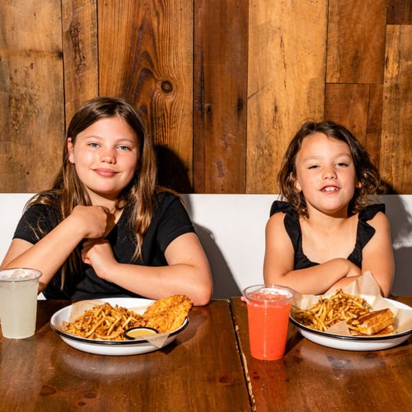 Two young girls smiling at a wooden table with plates of BuffBurger fries and chicken tenders.