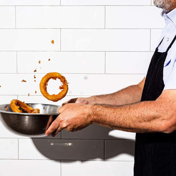 A chef in an apron tosses golden-brown BuffBurger onion rings from a metal bowl.
