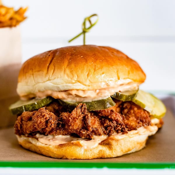 Crispy fried chicken sandwich with pickles and sauce on a toasted bun, with fries in the background.