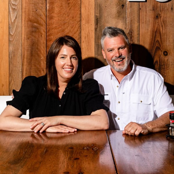 Two BuffBurger owners, a woman and a man, smiling at a wooden table with a wood-paneled wall.