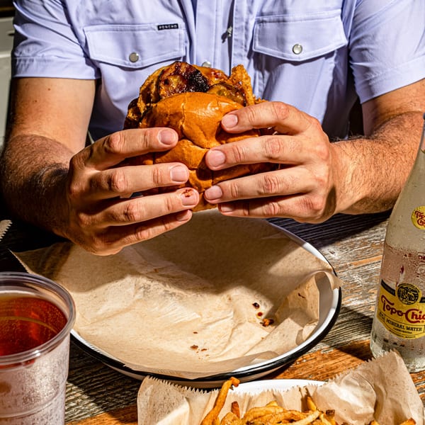 Hands holding a BuffBurger with a side of fries, a drink, and Topo Chico on a wooden table.