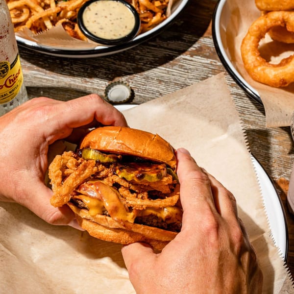 Hands holding a BuffBurger with crispy fried onions, pickles, and melted cheese, with fries and onion rings in the background.