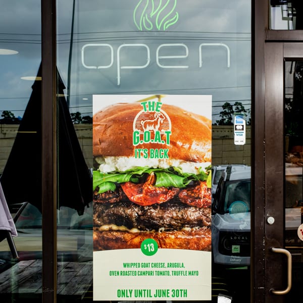 BuffBurger's "The G.O.A.T." burger advertisement on a glass door with an "open" neon sign.